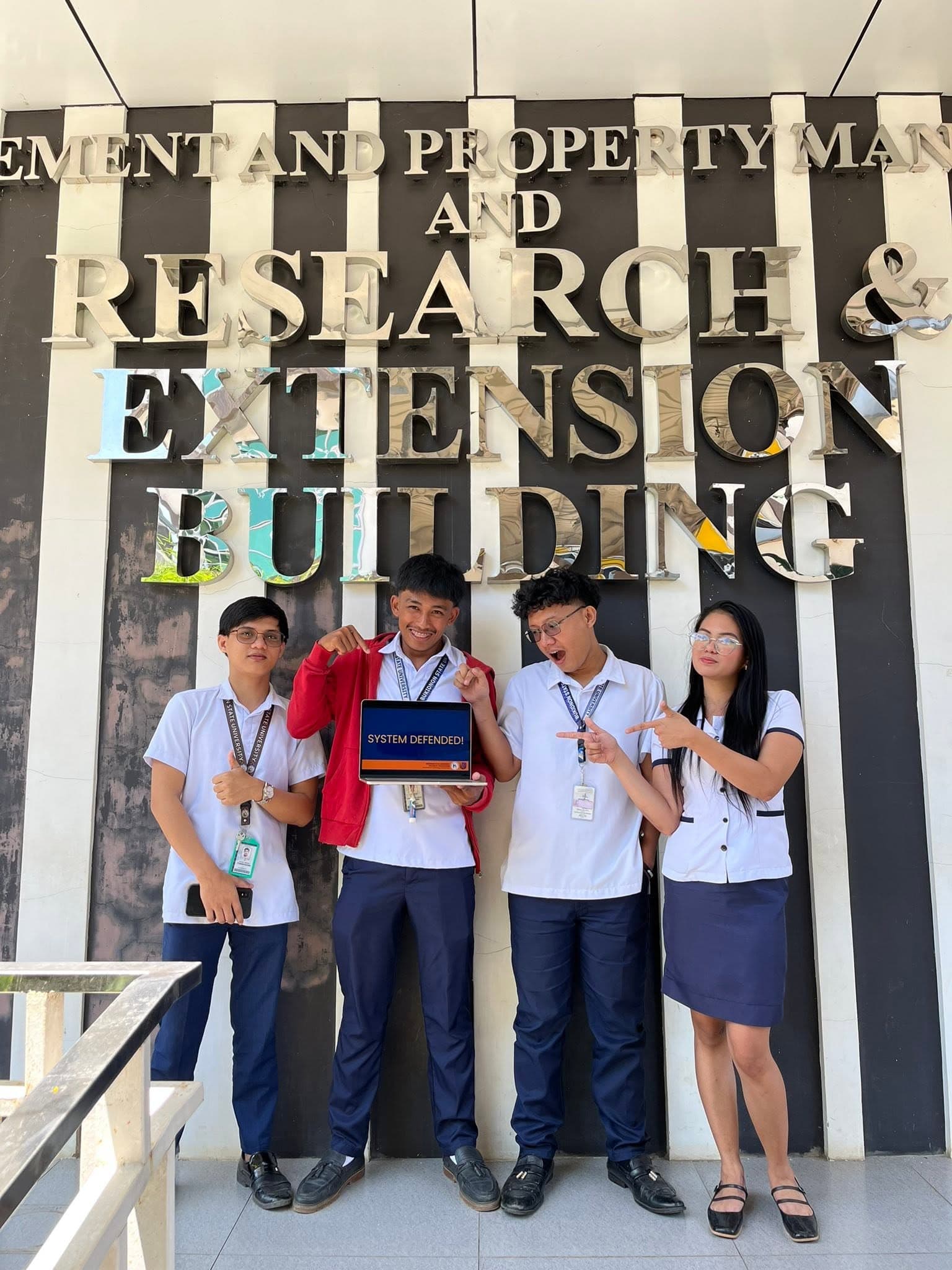 Proilan and groupmates holding a laptop showing "SYSTEM DEFENDED!" in front of the Bukidnon State University Research and Extension Building